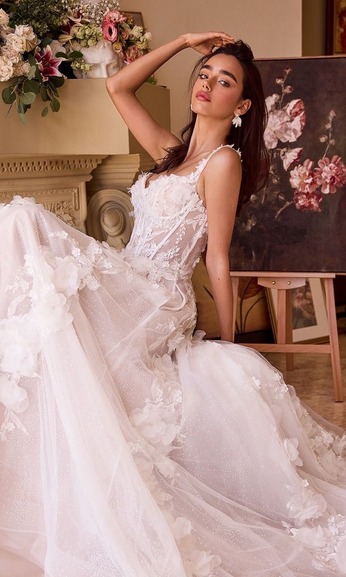 A woman wearing the Long Wedding Dress: Andrea and Leo WL045, featuring a lace corset bodice and tulle A-line skirt, sits on the floor beside a floral painting and a flower-adorned decorative mantle.